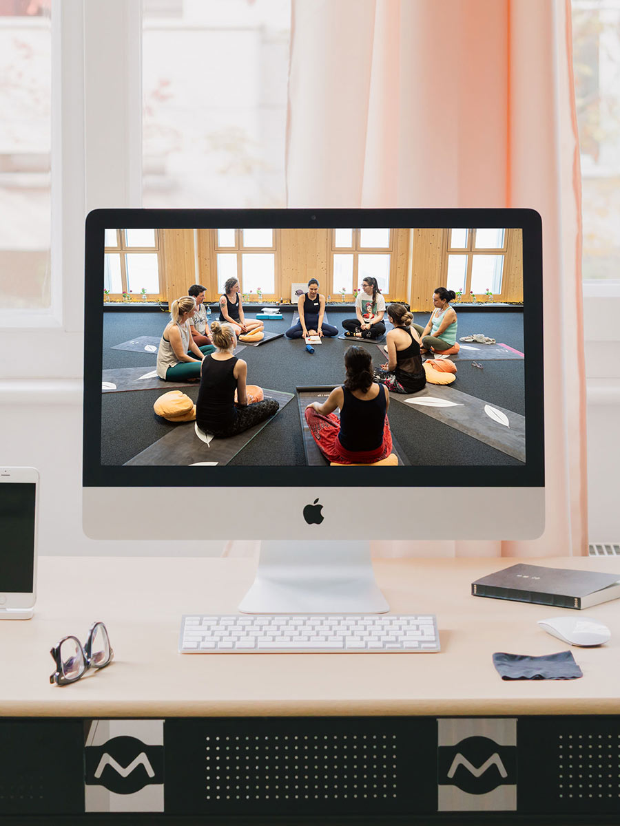 Auf einem Schreibtisch am Fenster steht ein großer Apple-Bildschirm, der Sam mit einer Yoga-Gruppe im Kreis sitzend zeigt.